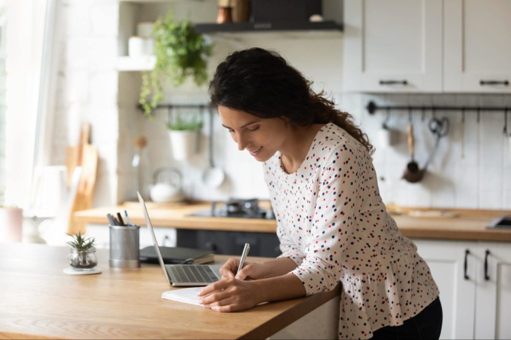 A picture of a woman in her kitchen creating a list with pen and paper.