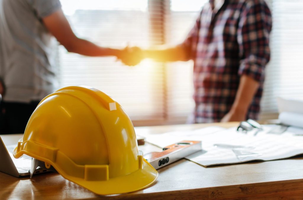 A close-up picture of a yellow hardhat with two men shaking hands in the background.