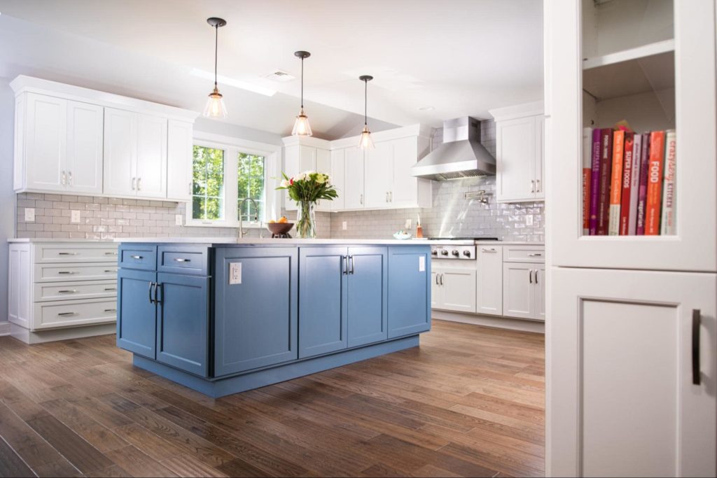 A picture of a modern kitchen with white cabinets and a large island in the middle with blue cabinets. On top of the island is a bowl of fruit and a vase with flowers in it.