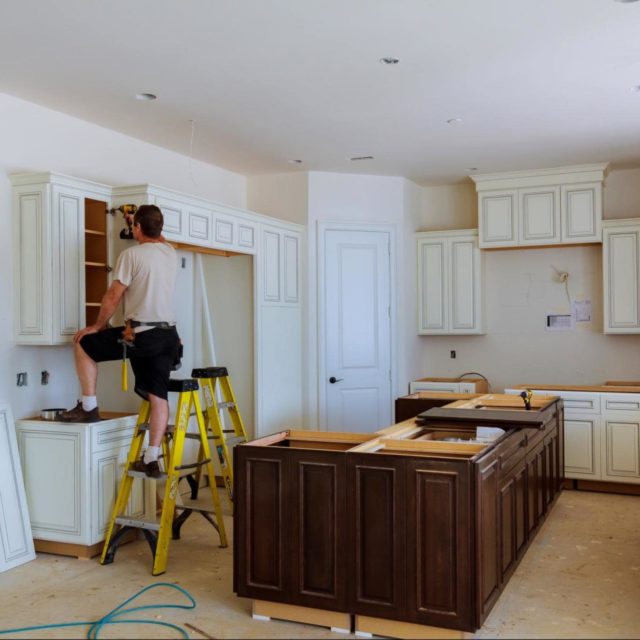 Contractor on a ladder drilling in a kitchen cabinet's doors.