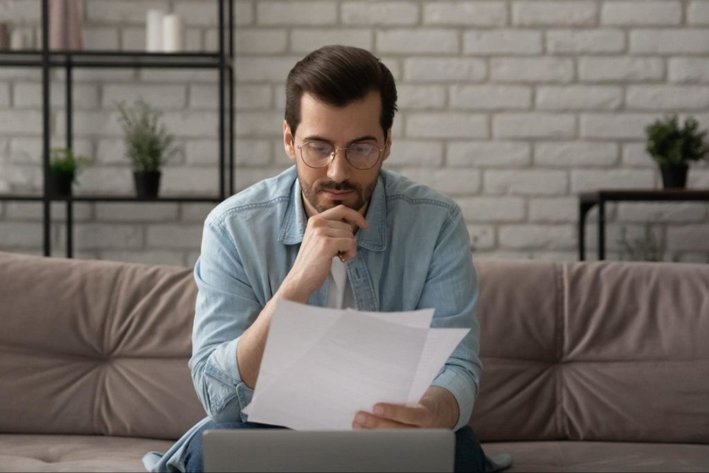 A young man sitting on his couch while looking over a contract with a laptop in front of him.