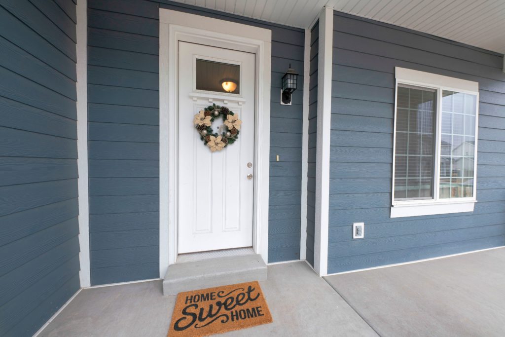 The white front door of a blue house with a rug that says "Home Sweet Home" on it.