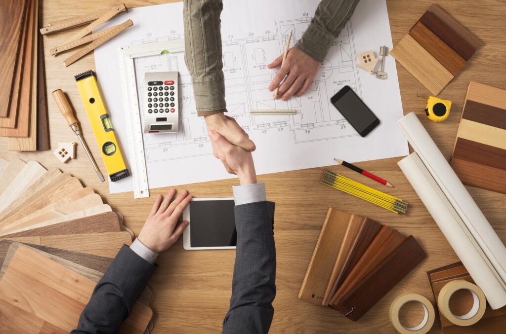 Two people shaking hands over building tools, blueprints, and wood samples.