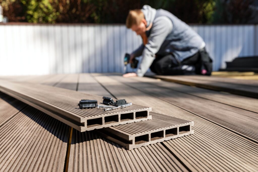 Decking materials sitting on a deck with a man using a drill in the background.