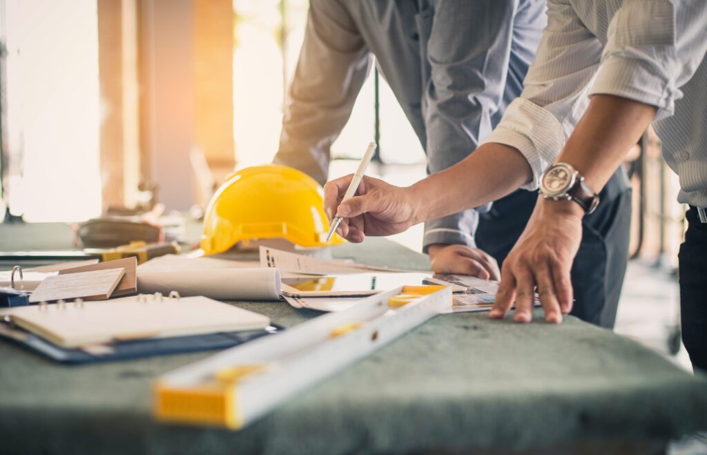 Two men standing over a drafting table at a construction site, going over paperwork and blueprints.