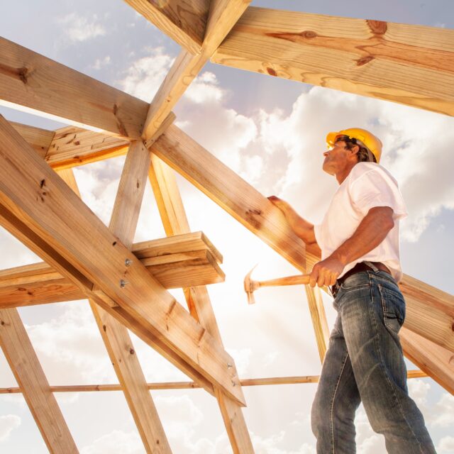 A construction worker standing under the framing for a roof in the sun.